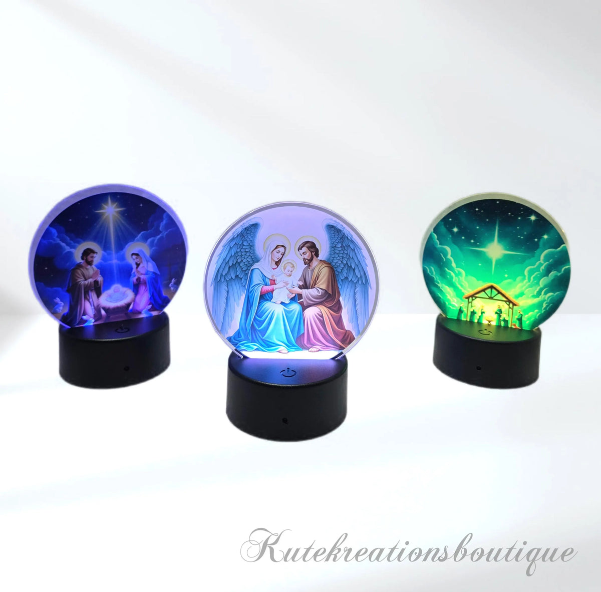 Three illuminated acrylic ball nativity lamps on black bases, shown side by side against a white background. The left lamp glows blue with a star and figures around a baby, the center lamp glows blue and pink showing Mary, Joseph, and baby Jesus, and the right lamp glows green with a stable scene under a bright star.